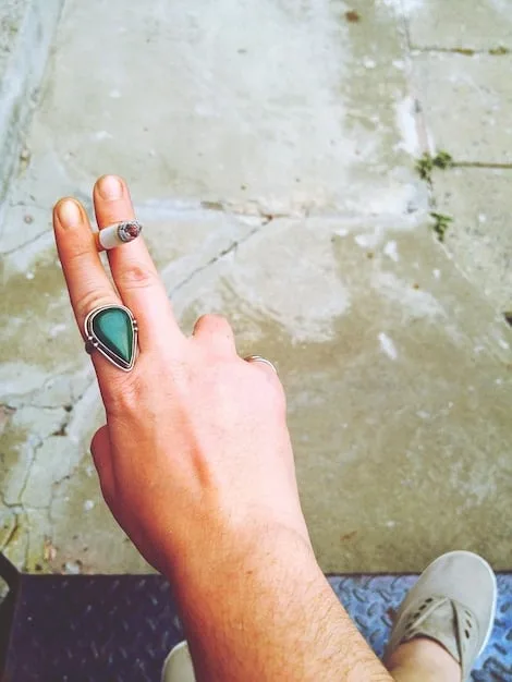 Close-up shot of a hand wearing multiple rings with different textures and natural stones, paired with a chunky resin bracelet. The background is a blurred image of a summer garden, emphasizing the nature-inspired theme.