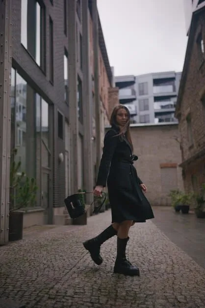 A woman wearing a beige trench coat over a simple black dress, accessorized with ankle boots and a crossbody bag, standing in an urban setting.