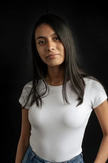 A close-up of a woman wearing a white crew neck t-shirt tucked into high-waisted black pants. She is accessorized with gold hoop earrings and a delicate gold necklace. The background is a blurred urban setting.