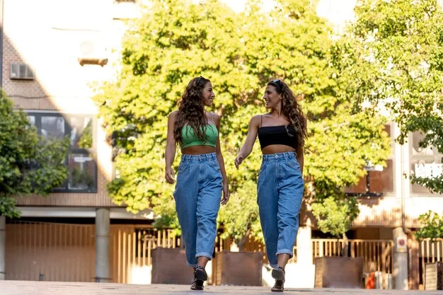Two women walking side-by-side on a sunny city street, both wearing 90s-inspired outfits. The first woman is wearing a plaid skirt and combat boots. The second is wearing black leggings under a sweatshirt, platform sneakers, and a backpack.