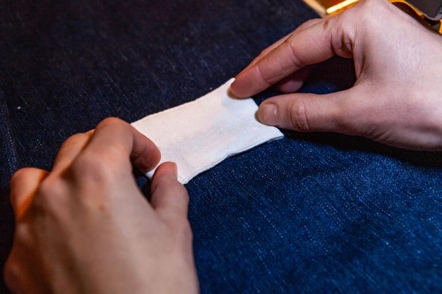 Close-up of a person's hands sewing a patch onto a denim jacket. The jacket is decorated with various patches and embroidery, showcasing a customized and upcycled look. The setting is a bright, cozy workspace.