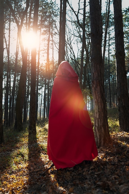 A woman standing in a forest, wearing a long, hooded cape in a deep burgundy color. The cape is made of wool and has a faux fur trim around the hood. The background is blurred, highlighting the cape and the woman's silhouette.