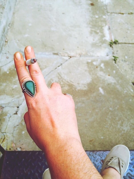 Close-up shot of a hand wearing multiple rings with different textures and natural stones, paired with a chunky resin bracelet. The background is a blurred image of a summer garden, emphasizing the nature-inspired theme.