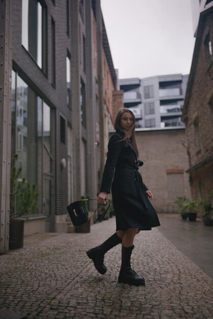 A woman wearing a beige trench coat over a simple black dress, accessorized with ankle boots and a crossbody bag, standing in an urban setting.