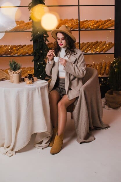 A woman wearing a long, elegant wool coat over a chic dress and high heels. She is standing in front of a softly lit restaurant, creating a warm and inviting atmosphere.